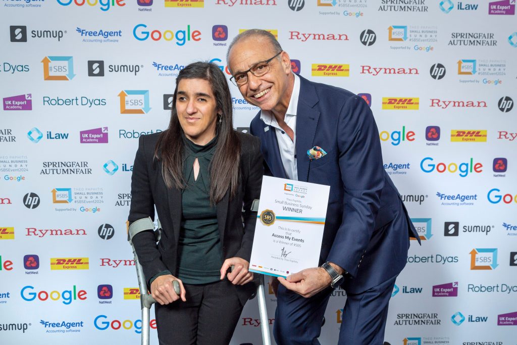 Access My Events founder, Zubee professional photo with Theo Paphitis. She is standing next to Theo on crutches and Theo is holding my certificate in his hand facing the camera in front, smiling. Behind them is a wall with logos of all the SBS partners and sponsors.