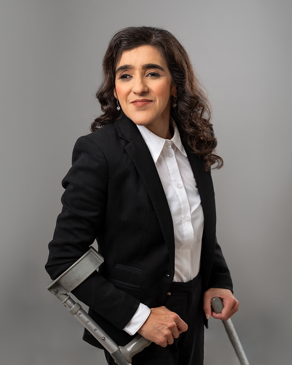 Zubee is standing with crutches and her head is turned slightly to the left looking at the camera, smiling. She is wearing a black blazer with a plain white collard shirt with closed buttons. She has curly hair opened with one side of her hair over her right shoulder