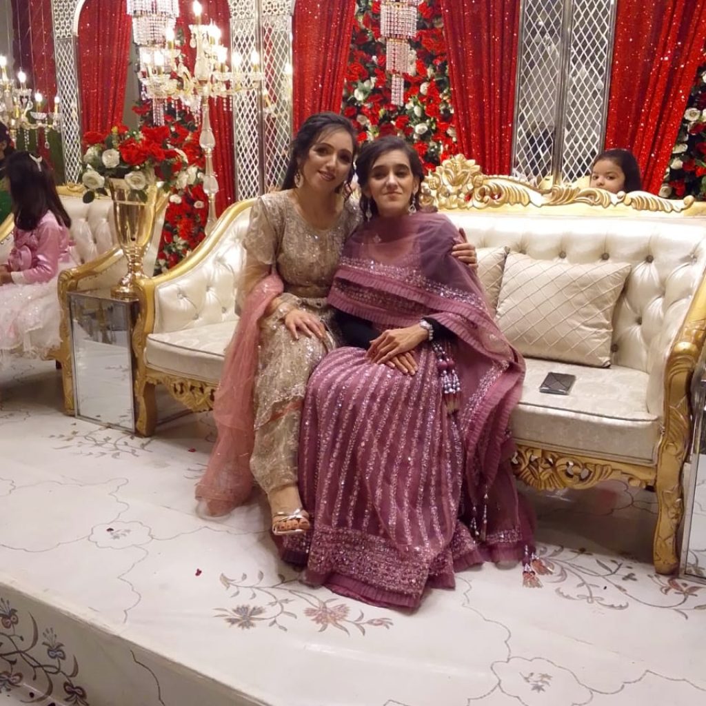 Zubee is sitting with her sister in a wedding hall on a long sofa that's set out on the stage where the bride and groom will sit. She is wearing her designer lengha by Oorvi Desai. It has silver embroidery lines going down the skirt with silver lines embroidery around the border of the skirt at the bottom. She also has a matching blouse at the top with a matching scarf, known as a duppata around her and a short black cardigan. Zubee's sister is sistting beside her wearing a beaded and sequin dress with a salwar (trouser) and a plain pink scarf. Behind them are red and white flowers decorated with big chandeliers coming down.
