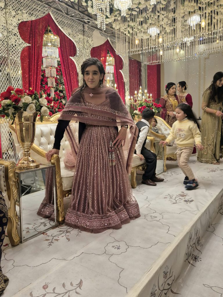 Zubee is standing holding on to a long sofa that's set out on the stage where the bride and groom will sit. She is wearing a designer lengha by Oorvi Desai. It has silver embroidery lines going down the skirt with silver lines embroidery around the border of the skirt at the bottom. She also has a matching blouse at the top with a matching scarf, known as a duppata around her and a short black cardigan. Behind her are red and white flowers decorated with big chandeliers coming down with some people and children playing.