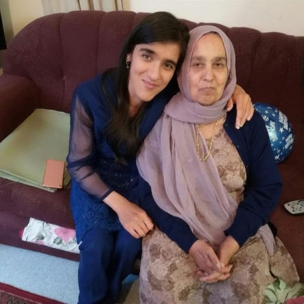 Zubee and her late grandmother sitting on the sofa next to each other with Zubee's arm around her nan looking in front slightly smiling together. We are South Asian wearing our traditional outfits a salwar, known as a trouser and a kameez known as a short dress. Zubee is wearing a royal blue colour and her grandmother is wearing a light pinky/beige with printed flowers. 
