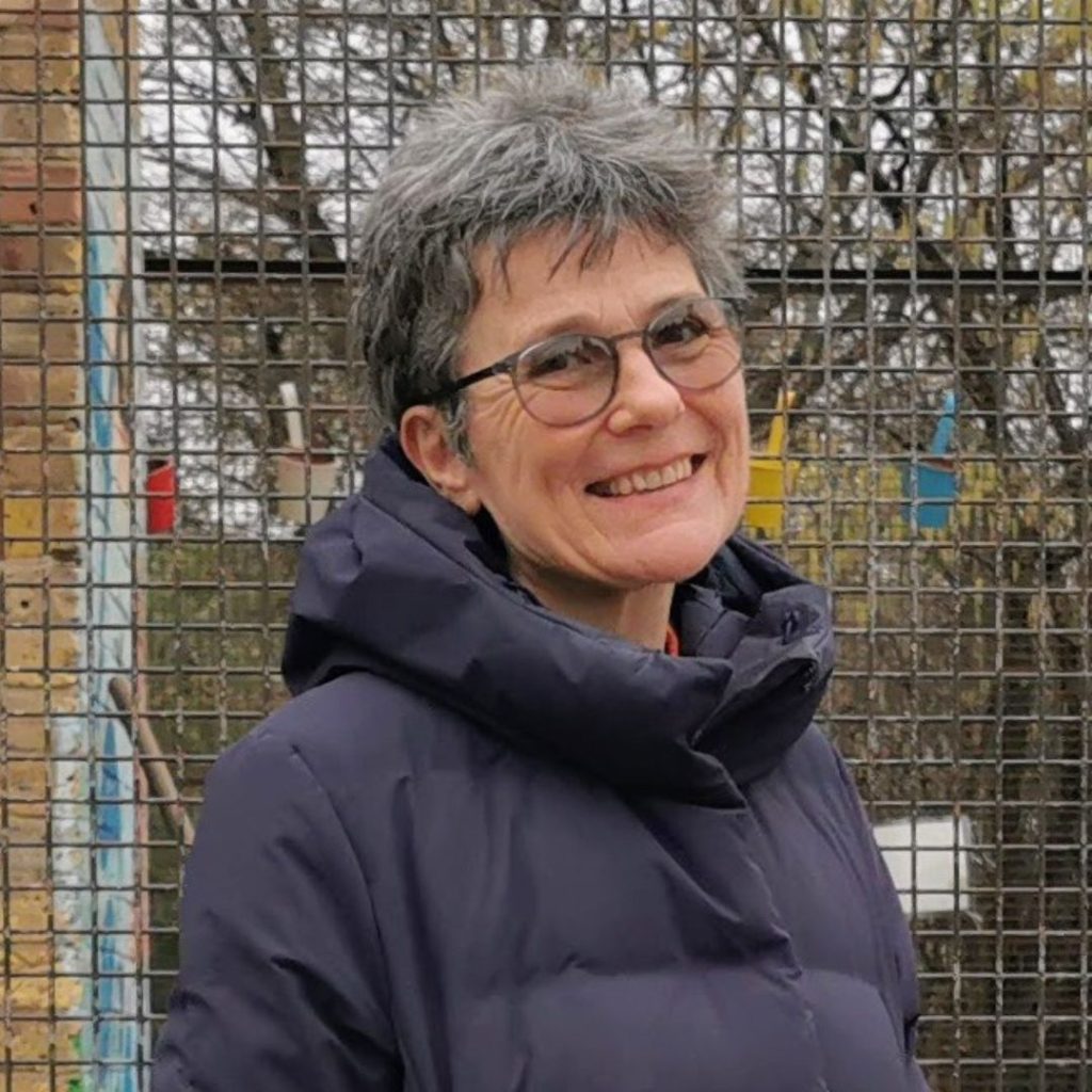 Clare, the CEO of For Brian is standing in the outdoors with a fence behind her. She has grey short hair with glasses and is wearing a blue long puffer coat, looking in front smiling. 