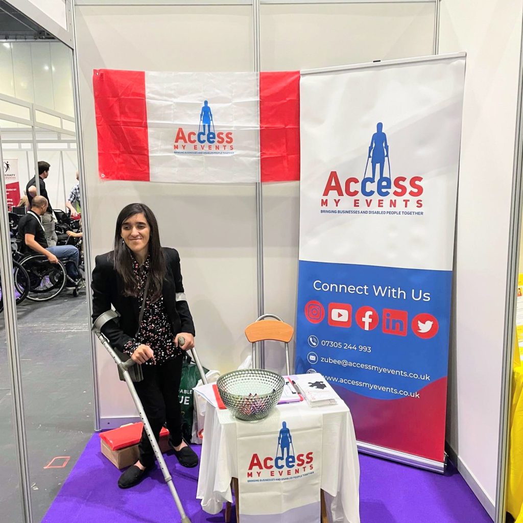 An indoor photo shows Zubee standing at a trade show at her  stall for "Access My Events." She is wearing a black blazer and patterned top, and she is using a forearm crutches. The stand features a red and white flag, a tall banner, and a small table covered with a white cloth, all displaying the "Access My Events" logo, which shows a silhioutte of a person using crutches. The banner also includes social media icons and contact information. The stall is set on a purple carpet. In the background, other people and event displays are visible.
