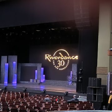 At the Eventim Apollo Hammersmith. In front is the stage with a lot of empty chairs for audience members as they arrive. There is purple coloured lighting and a few steps on the stage. There is a black backdrop that says Riverdance 30 The New Generation in gold text in a circle.