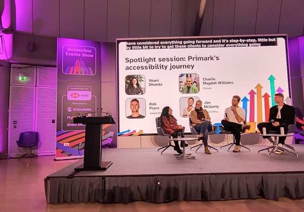 3 panellists sitting on chairs on stage with host Shani asking questions. Behind them is a screen that reads spotlight session Primark's accessibility journey with images and names of each panellist. On top, it shows live captions.