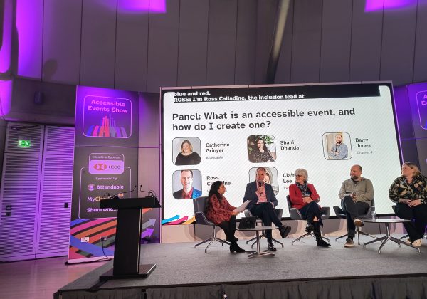 4 panellists sitting on chairs on stage with host Shani asking questions. Behind them is a screen that reads Panel: What is an accessible event, and how do I create one? with images and names of each panellist. On top, it shows live captions.