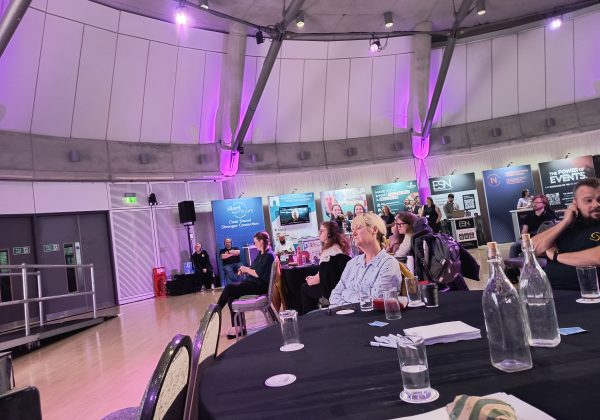A round table in front with audience members sat around tables facing the stage. Further back is a display of a few stalls and company banners.