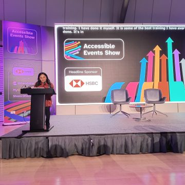 Shani, is one of the organisers of the event.  She is South Asian with below-the-shoulder black length curly hair. She is standing on stage facing the audience with a podium and microphone in front of her. Behind her is a large screen with the accessible events show logo on a black background with a logo of the headline sponsor, HSBC and a banner displaying the same info next to the screen. There is a small round table with two chairs side by side on the stage.