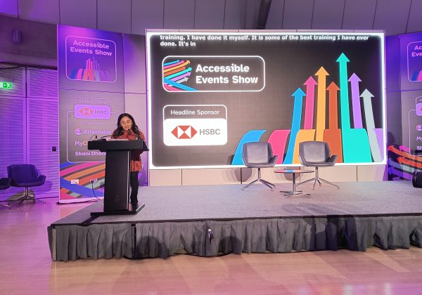 Shani, is one of the organisers of the event.  She is South Asian with below-the-shoulder black length curly hair. She is standing on stage facing the audience with a podium and microphone in front of her. Behind her is a large screen with the accessible events show logo on a black background with a logo of the headline sponsor, HSBC and a banner displaying the same info next to the screen. There is a small round table with two chairs side by side on the stage.