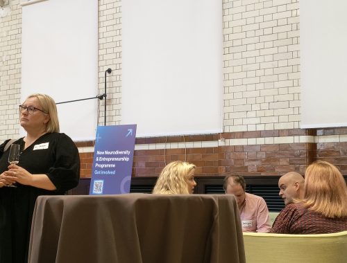 A few attendees sitting on chairs chatting with a round table away from them. Next to them is a banner that says new neurodiversity  and entrepreneurship programme get involved with a QR code below. An attendee is next to the table and has a drink in her hand.