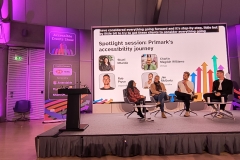 3 panellists sitting on chairs on stage with host Shani asking questions. Behind them is a screen that reads spotlight session Primark's accessibility journey with images and names of each panellist. On top, it shows live captions.