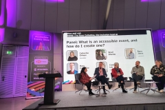4 panellists sitting on chairs on stage with host Shani asking questions. Behind them is a screen that reads Panel: What is an accessible event, and how do I create one? with images and names of each panellist. On top, it shows live captions.