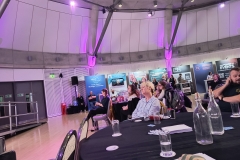 A round table in front with audience members sat around tables facing the stage. Further back is a display of a few stalls and company banners.