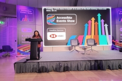 Shani, is one of the organisers of the event.  She is South Asian with below-the-shoulder black length curly hair. She is standing on stage facing the audience with a podium and microphone in front of her. Behind her is a large screen with the accessible events show logo on a black background with a logo of the headline sponsor, HSBC and a banner displaying the same info next to the screen. There is a small round table with two chairs side by side on the stage.