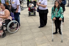 Zubee is standing looking in front of the camra smiling. She is standing with crutches and there is a small crowd of wheelchair users and people watching 