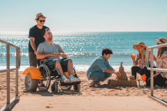 An image of a sandy beach with the sun shining on it. There is a wheelchair user on the sand, smiling with a guy holding the handles of his chair from behind him and is smiling. There are people sitting next to them with drinks in their hands and a boy is building a sand castle  in front of them