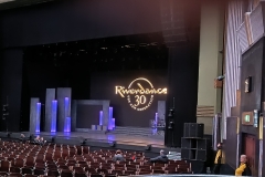 At the Eventim Apollo Hammersmith. In front is the stage with a lot of empty chairs for audience members as they arrive. There is purple coloured lighting and a few steps on the stage. There is a black backdrop that says Riverdance 30 The New Generation in gold text in a circle.