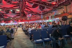 Audience SBS attendees sitting down in the conference hall, which is slowly filling up as people are coming in waiting for the event to start. On the ceiling are white drapes with banners against the walls with a reflection of red light.