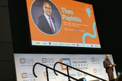 Theo Paphitis on stage. Behind him is a screen that shows an image of Theo, his title and his social media handles on X and LinkedIn, with the SBS banner below.