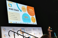 Theo is on stage facing the audience. The TV screen displayed on the wall behind Theo that reads #SBS is 15 showing some stats over the years since SBS started Below the screen is a big banner that reads Small Business Sunday supported by Google with their logo below the SBS logo on a white background.