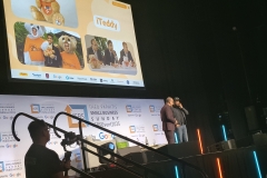 Theo and Imran on stage. The screen behind them shows the iTeddy and images of Theo and Imran sitting at a table with another image showing his family.   Below the TV screen is the SBS banner that says Theo Paphitis Small Business Sunday supported by Google with their logo on a white background.