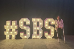 Access My Events founder, Zubee is standing on crutches next to neon letters that say hashtag SBS   that are lit up in big gold letters