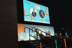 Theo and Firside chat guest with Richard Harpin on stage. The screen behind them shows an image of Richard and Theo with their names below. Theo and Richard are sitting facing each other on stage